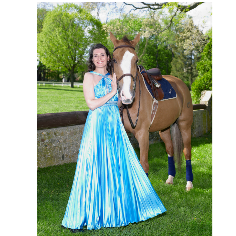 Silvana Dugandžić Penavić, founder of Raul E® New York, in a flowing blue pleated gown standing beside her horse, Raul